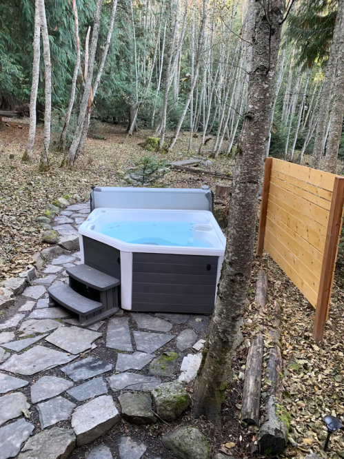 A hot tub on a stone path surrounded by trees and autumn leaves, with a wooden fence in the background.