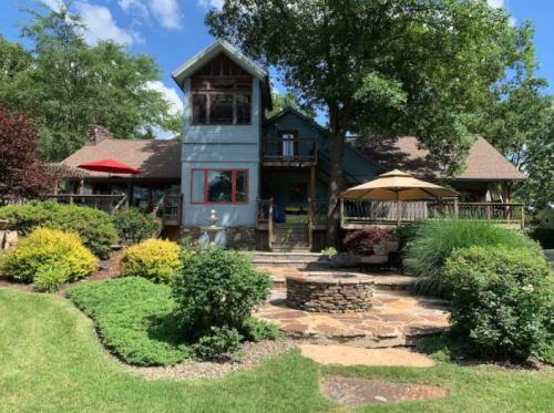 A charming house with a stone patio, surrounded by lush greenery and trees, under a clear blue sky.