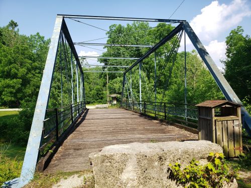 A blue metal truss bridge spans a small area, surrounded by lush green trees and a wooden structure nearby.