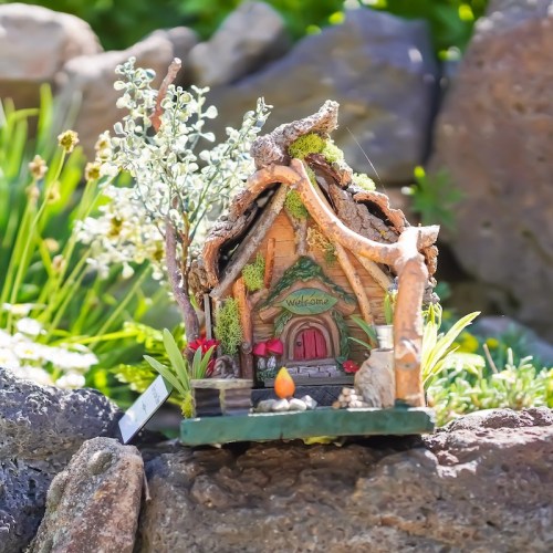 A whimsical miniature cottage surrounded by greenery and flowers, with a "Welcome" sign at the entrance.
