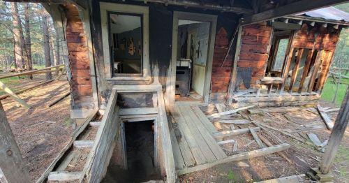 An abandoned, weathered cabin with peeling paint and a broken porch, surrounded by trees and overgrown vegetation.