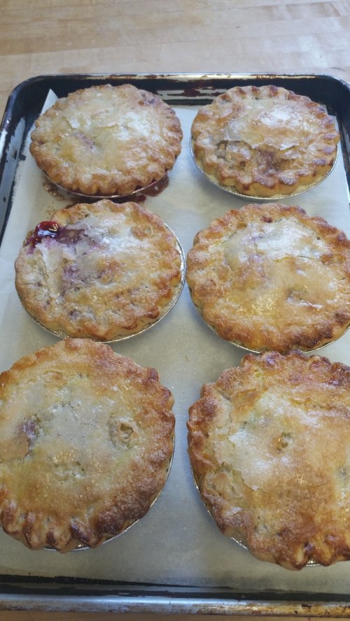 Six golden-brown mini pies on a baking tray, with a flaky crust and a hint of fruit filling peeking out.