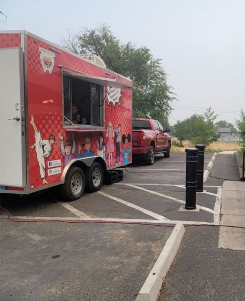 A colorful food truck parked in a lot, with a red pickup truck waiting in line nearby. Trees in the background.