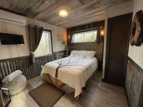 Cozy bedroom with a tree-patterned bedspread, wooden walls, and natural light from windows.