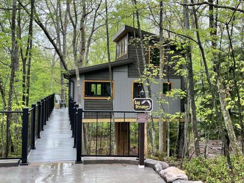 A modern treehouse nestled in a forest, featuring large windows and a walkway leading up to it.