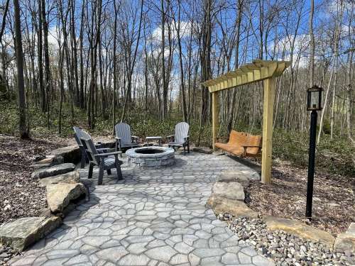 A serene outdoor seating area with a fire pit, stone path, and wooden swing, surrounded by trees and blue sky.