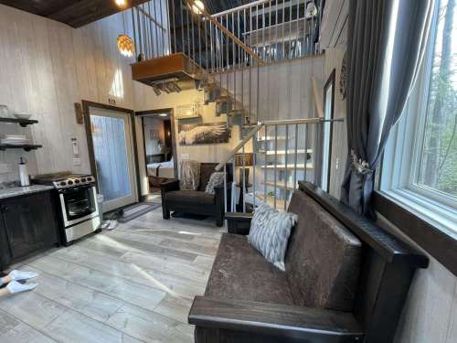 Cozy interior of a cabin featuring a kitchen, seating area, and staircase leading to an upper level. Natural light streams in.