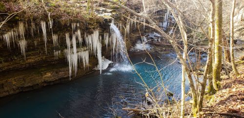 A serene waterfall cascades into a blue pool, surrounded by icy formations and bare trees in a tranquil forest setting.
