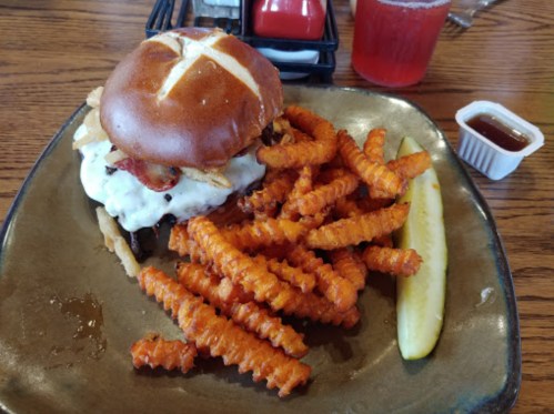 A pretzel bun burger topped with bacon and sauce, served with crinkle-cut sweet potato fries and a pickle slice.
