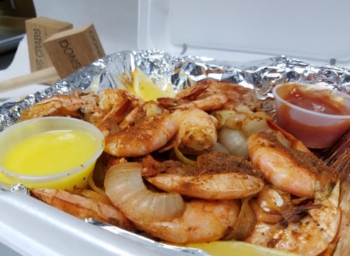A foil tray filled with seasoned shrimp, onions, lemon wedges, and two dipping sauces.