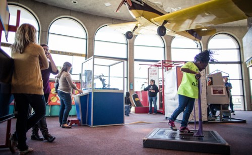 A child in a bright yellow shirt plays with an interactive exhibit in a lively science museum filled with visitors.