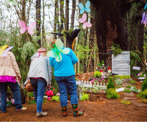 Three people with butterfly wings explore a whimsical garden decorated with flowers and fairy-themed elements in a forest.