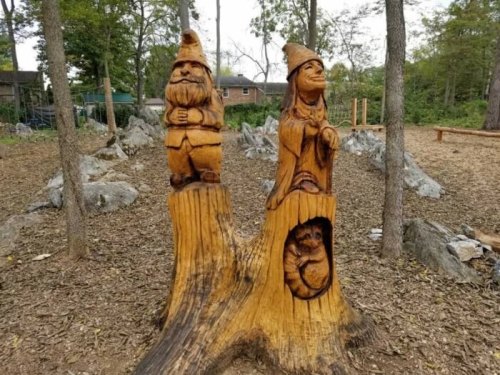 Wooden carvings of a gnome and a woman, with a small animal peeking out, set in a natural outdoor environment.