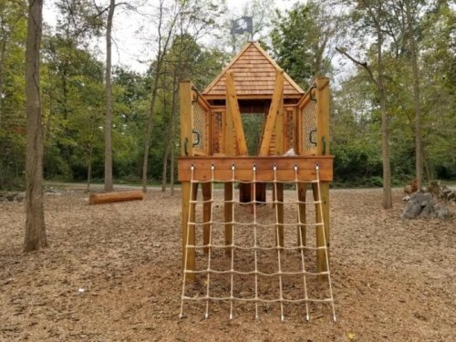 A wooden pirate-themed play structure with a climbing net, surrounded by trees and a sandy ground.
