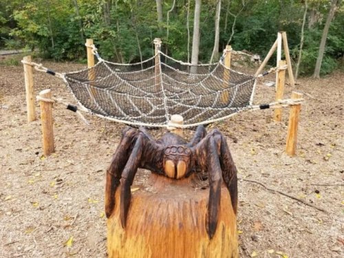 A wooden spider sculpture sits on a stump near a rope web climbing structure in a forested area.