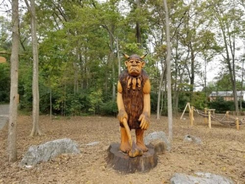 A wooden sculpture of a troll stands in a forested area, surrounded by trees and rocks.