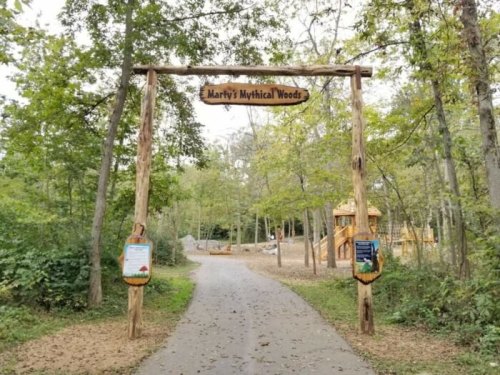 Wooden archway sign reading "Marty's Mythical Woods" at the entrance of a forested path with trees and play structures.