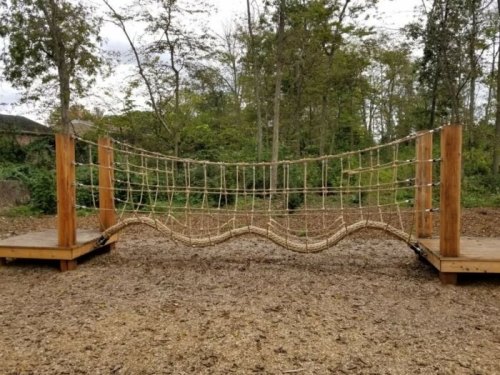 A rope bridge with wooden supports, set in a forested area with trees in the background.