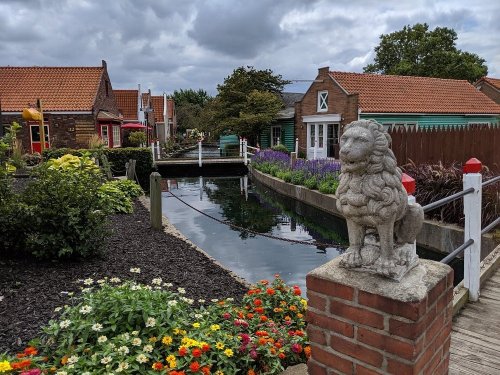 A quaint village scene with colorful houses, a canal, and a stone lion statue surrounded by flowers.