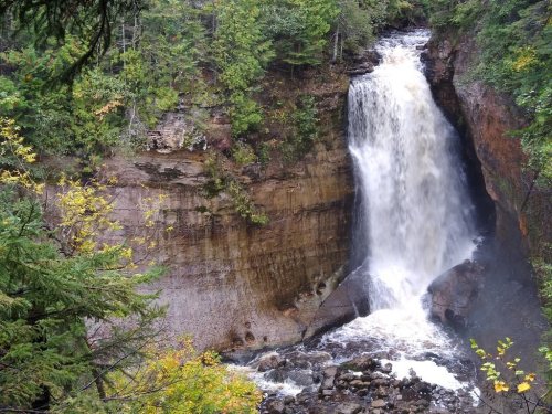 A cascading waterfall surrounded by lush green trees and rocky terrain, creating a serene natural scene.