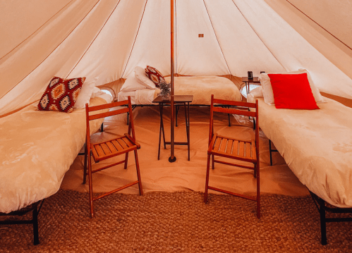 Cozy glamping tent with two beds, wooden chairs, and a small table, featuring warm, inviting decor.