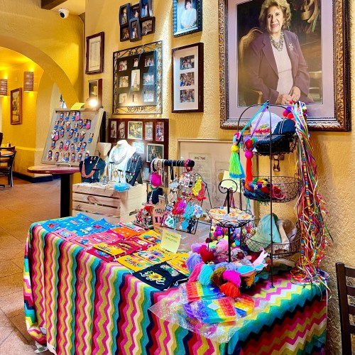 Colorful display of handmade crafts and jewelry on a table, with framed photos and a portrait on the wall behind.