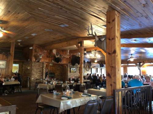 Cozy restaurant interior with wooden beams, mounted animal heads, and diners at tables. Warm, rustic atmosphere.