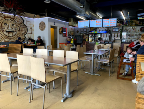 A spacious restaurant interior with wooden tables and chairs, featuring a child in a high chair and patrons in the background.