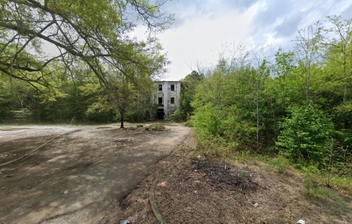 Abandoned building surrounded by overgrown trees and shrubs, with a gravel path leading towards it.