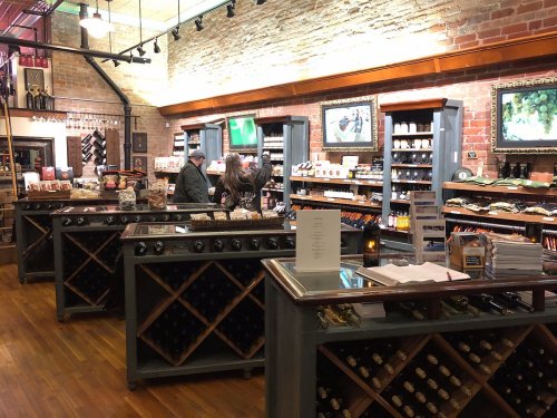 A cozy wine shop with wooden shelves, brick walls, and customers browsing various wine and gourmet food products.
