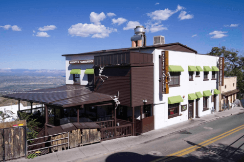A two-story restaurant with a brown exterior and green awnings, set against a scenic mountain backdrop and blue sky.