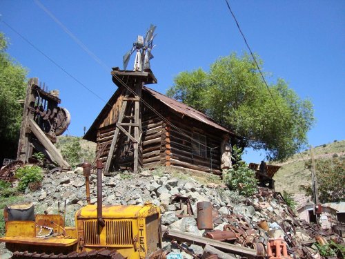 A rustic log cabin with a windmill, surrounded by old machinery and greenery under a clear blue sky.