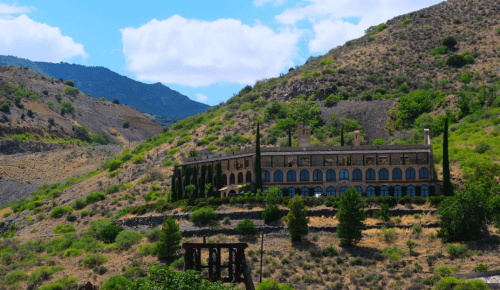 A historic building nestled on a hillside, surrounded by greenery and mountains under a blue sky with clouds.