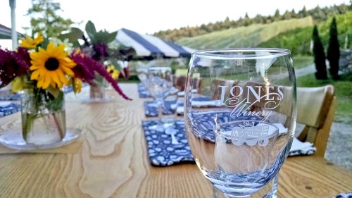 A beautifully set outdoor dining table with a glass featuring "Jones Winery" and vibrant flowers in the background.