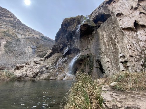 A serene waterfall cascades down rocky cliffs into a calm pool, surrounded by dry grass and rugged terrain.