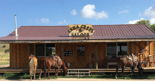 A rustic wooden saloon with a metal roof, featuring three horses tied up in front under a blue sky.