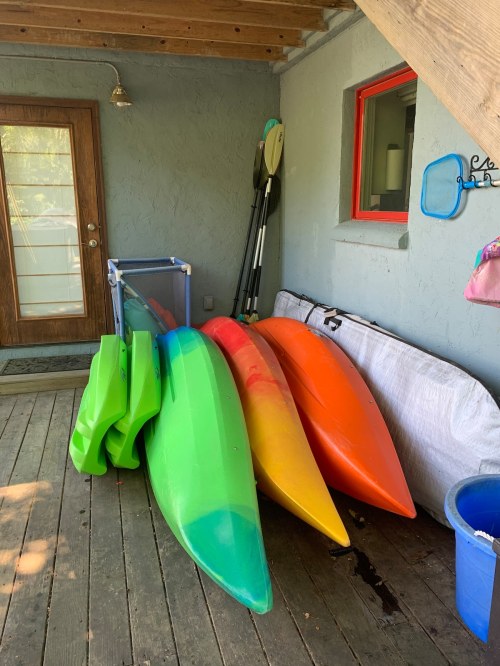 Colorful kayaks in green, orange, and yellow stacked against a wall on a wooden deck, with paddles and a blue bin nearby.