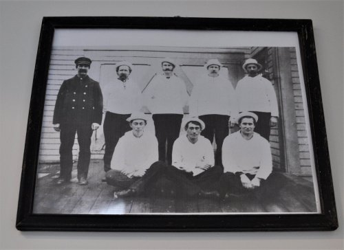 Black and white photo of nine men in sailor attire, posed indoors, with one man in a captain's uniform.