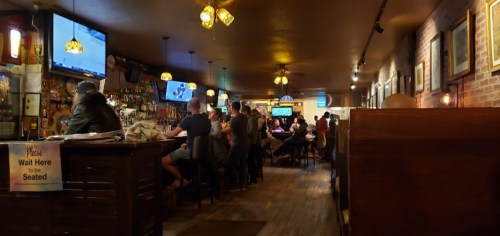 A lively bar scene with patrons at the counter and tables, featuring TVs and warm lighting.
