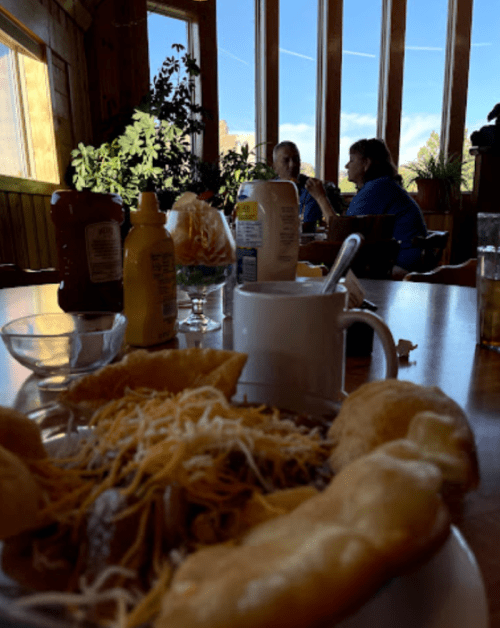 A cozy restaurant scene with a plate of food in the foreground and two people chatting by a window.