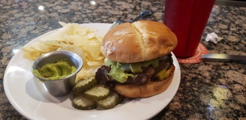 A burger with guacamole, pickles, and potato chips on a plate, with a red drink in the background.