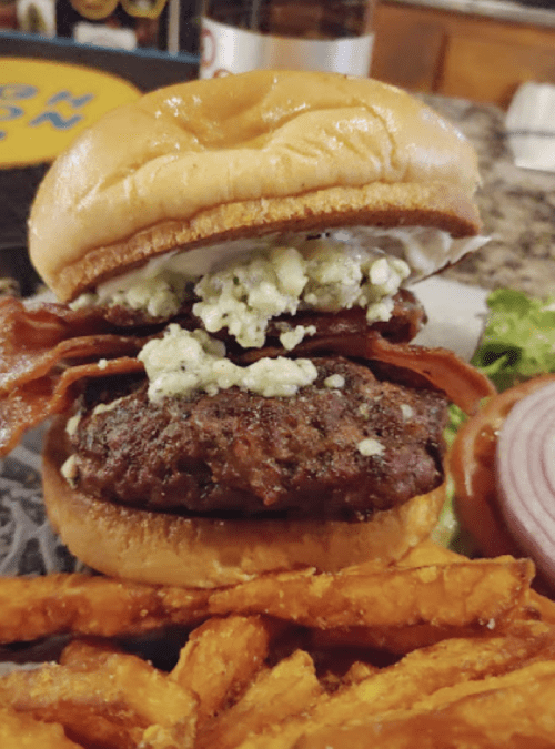 A juicy burger with blue cheese, bacon, and lettuce on a bun, served with sweet potato fries on the side.