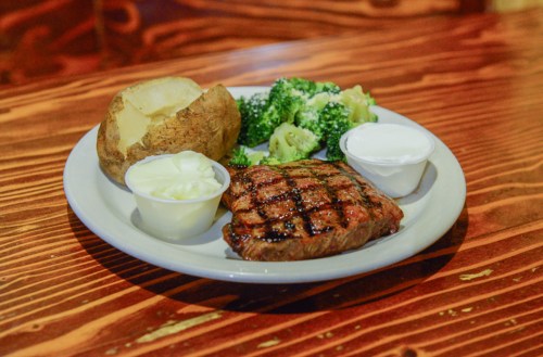 A plate with a grilled steak, baked potato, steamed broccoli, and two cups of dipping sauce on a wooden table.