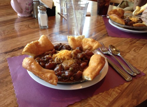 A plate of chili topped with cheese, surrounded by fried dough, on a wooden table with utensils and drinks in the background.