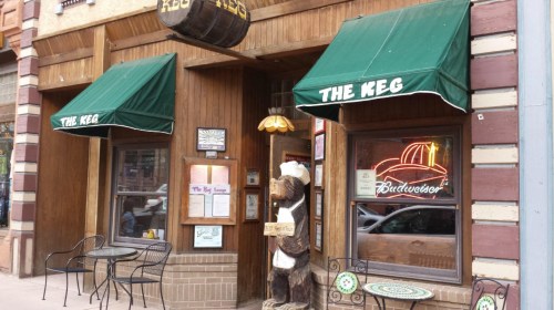 Exterior of a rustic restaurant called "The Keg," featuring green awnings, wooden decor, and a bear statue.