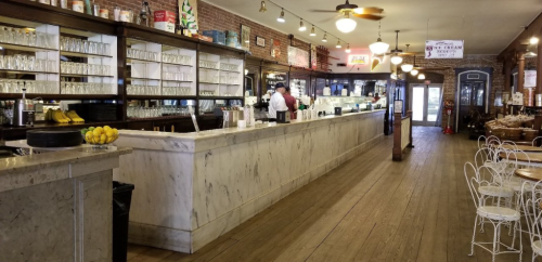 A vintage ice cream parlor with a marble counter, glassware, and wooden decor, featuring tables and chairs.