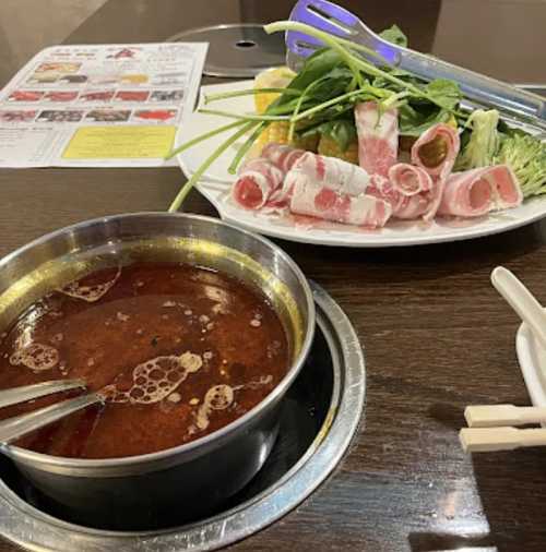 A hot pot with broth and raw meat slices, accompanied by fresh vegetables on a plate, set on a wooden table.