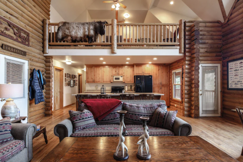 Cozy log cabin interior featuring a living area, kitchen, and a loft with a decorative cow on the railing.