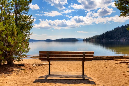 A wooden bench on a sandy shore overlooking a calm lake, surrounded by trees and mountains under a blue sky.