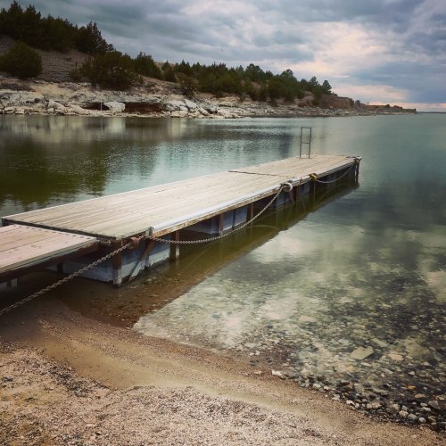 A wooden dock extends into calm water, surrounded by a rocky shoreline and trees under a cloudy sky.